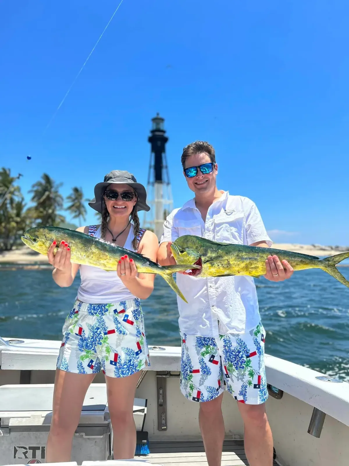 A couple holding up two mahi mahi with the Hillsboro Inlet lighthouse behind them
